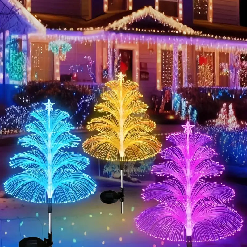 Three illuminated tree-shaped fiber optic solar lights in blue, yellow, and pink colors decorating a house lawn for the holidays.