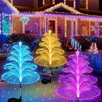 Three illuminated tree-shaped fiber optic solar lights in blue, yellow, and pink colors decorating a house lawn for the holidays.